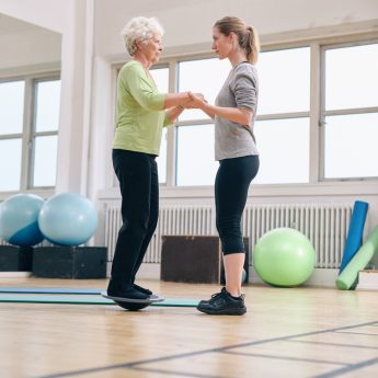 A woman helping an older woman standing on a balance board.